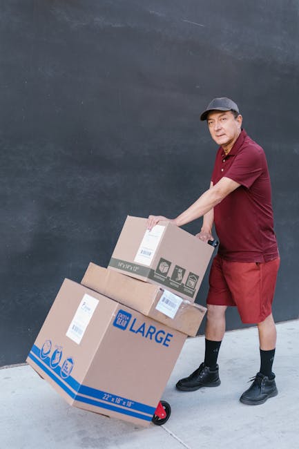 A delivery person wearing a burgundy shirt, matching shorts, black socks, and black shoes is seen pushing a hand truck loaded with three brown cardboard boxes, one labeled 'LARGE' and with UK courier 