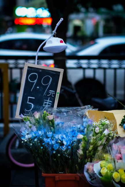 A bouquet of fresh blue and pale pink flowers, possibly including miniature roses and filler blooms, arranged in clear plastic wrapping and placed in front of a chalkboard sign displaying prices in Ch