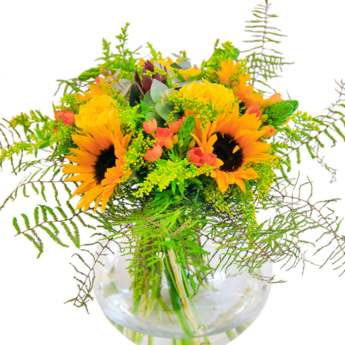 This bouquet, artfully crafted by a skilled Crouch End florist, evokes the golden heart of summer with an abundant collection of radiant sunflowers, their bright petals forming luminous halos around velvety, deep brown centers. Alongside, opulent yellow garden roses unfurl in lush spirals, each petal gently layered, exuding classic elegance reminiscent of the villa gardens found on Tottenham Lane. Glossy clusters of red Hypericum berries nestle amid the vivid blooms, lending spirited pops of colour that spark against the warm yellows and rich greens. Delicate fern fronds cascade over the arrangement's edges, mimicking the undulating greenery of local woodland walks, while dainty stems of yellow solidago and ethereal blue-green eucalyptus add lightness and aromatic freshness, reminiscent of a Sunday at Stationers Park. All are gathered in a clear, globe-shaped glass vase, its surface gleaming under natural light and its depths revealing the intertwining stems and the pure water that keeps every flower at peak vibrancy. Up close, layers of texture become apparent: velvety sunflower disks, plush rose petals, the waxy sheen of berries, and the crisp snap of fresh foliage-all combining in a harmonious interplay that feels at once wild and artful. The bouquet's soft, sweet scent drifts gently, evoking feelings of open fields and late afternoon sun, making it an ideal centrepiece for family gatherings, celebrations, or moments of gratitude. This composition honours both the artistry and the natural abundance found right here in Crouch End.